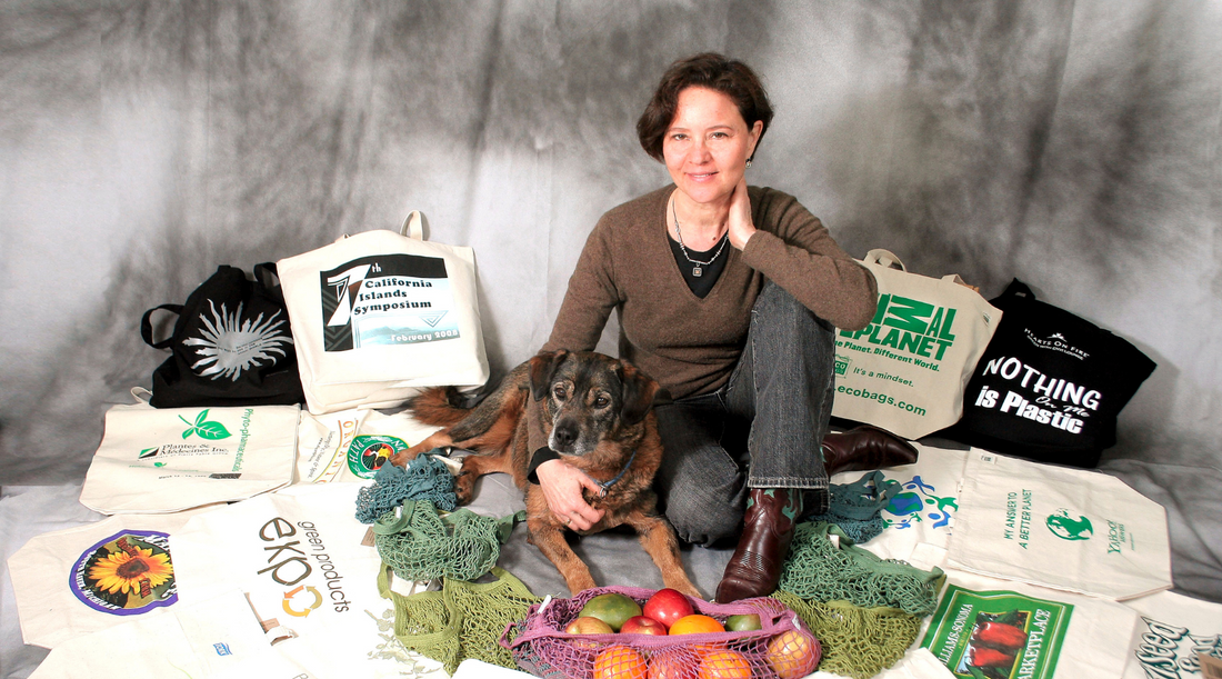 Sharon Rowe, a woman with short brown hair and a light complexion, sits among an array of printed ECOBAGS totes and string bags with her brown dog, Hudson.