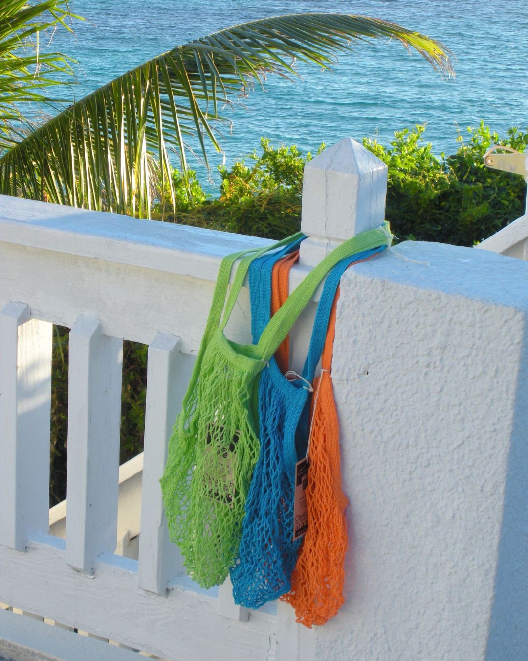 Three mesh reusable shopping bags in green, blue, and orange hanging on a white railing with a scenic tropical background.