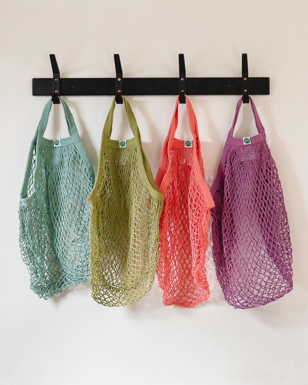 Four colorful mesh net bags hanging on a black rod against a white wall with a visible green logo.