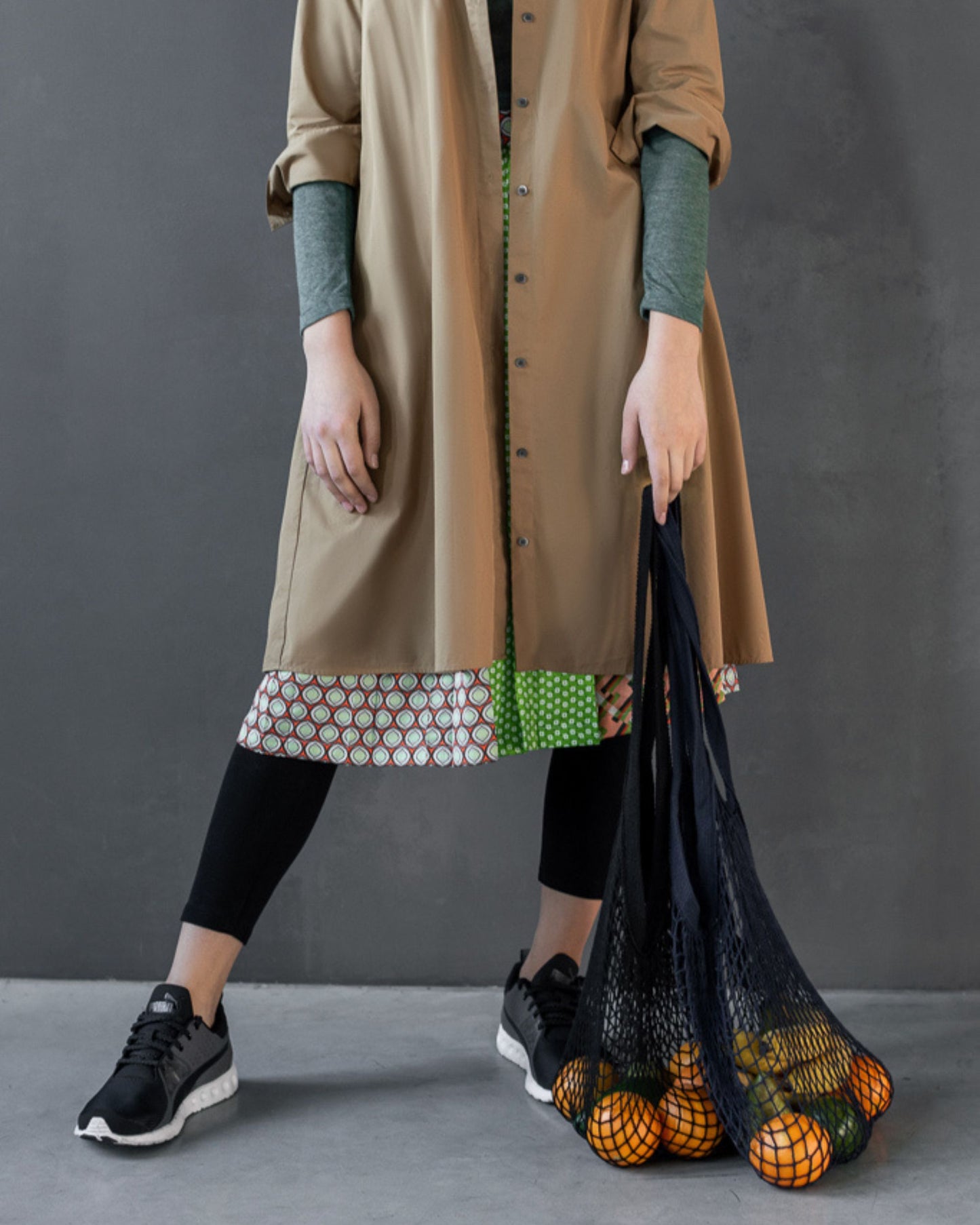 Person wearing a long beige coat with patterned hem, holding a black french market string bag with oranges against a gray background.