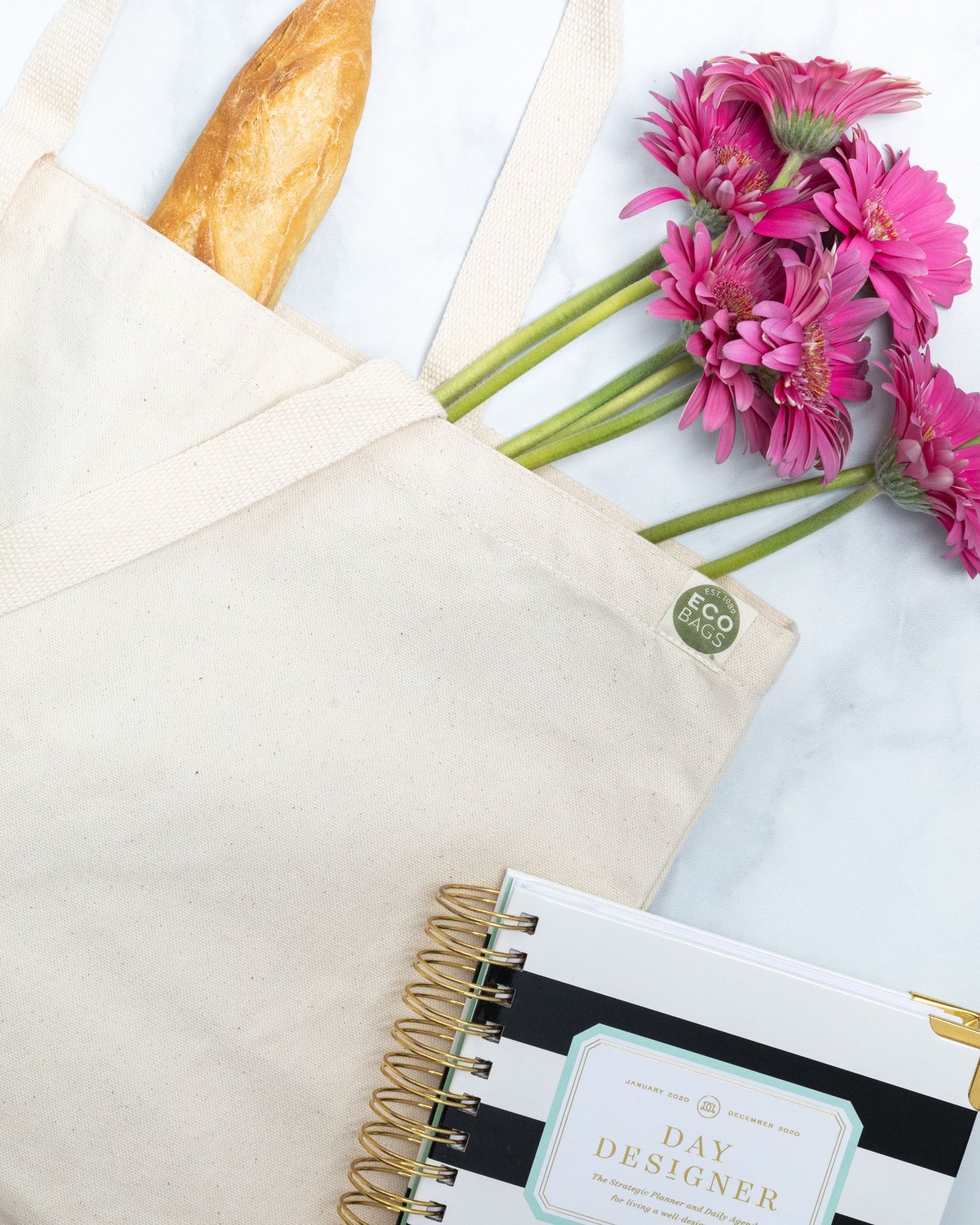 Natural colored recycled cotton tote bag with a loaf of bread, pink flowers, and a notebook on a light background