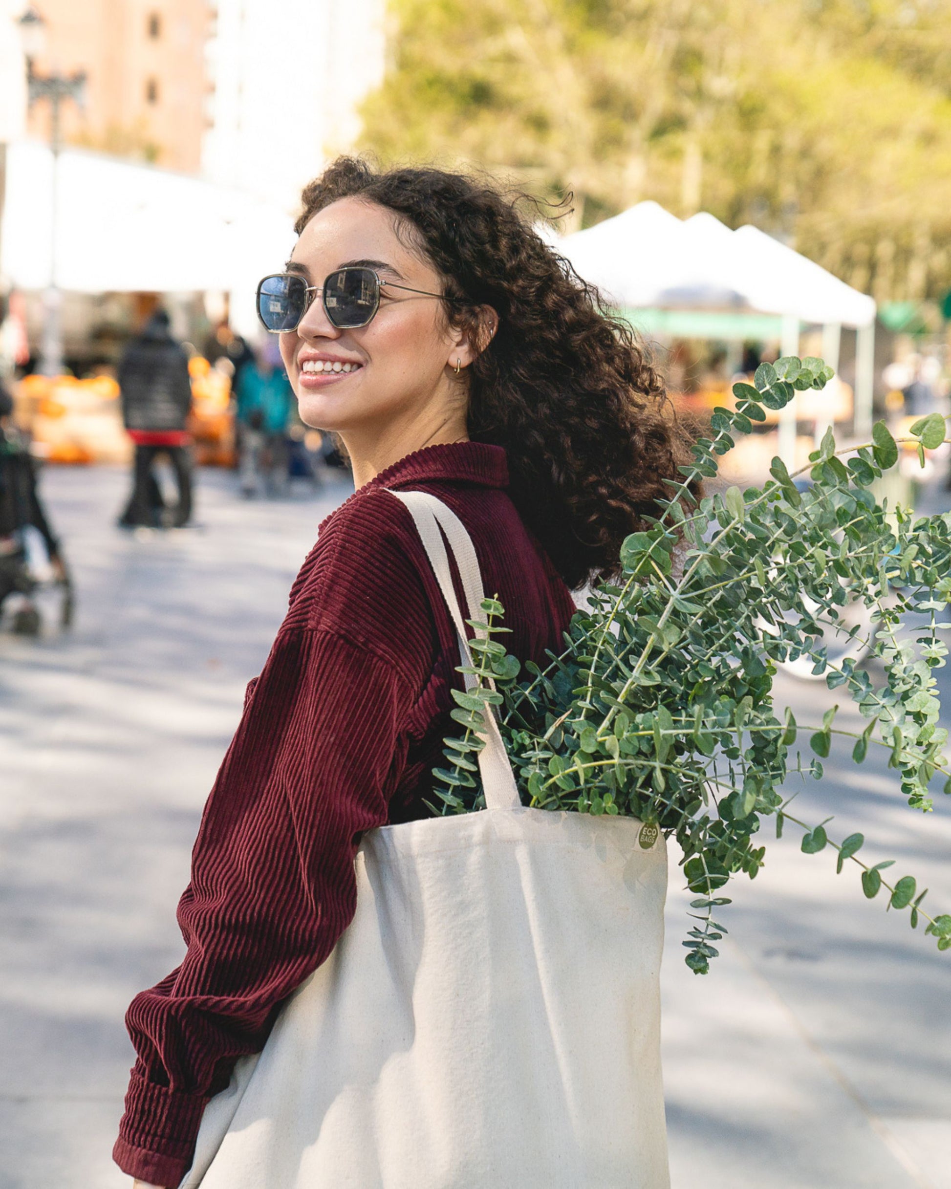 Woman with sunglasses and a natural canvas tote bag holding plants in an outdoor setting