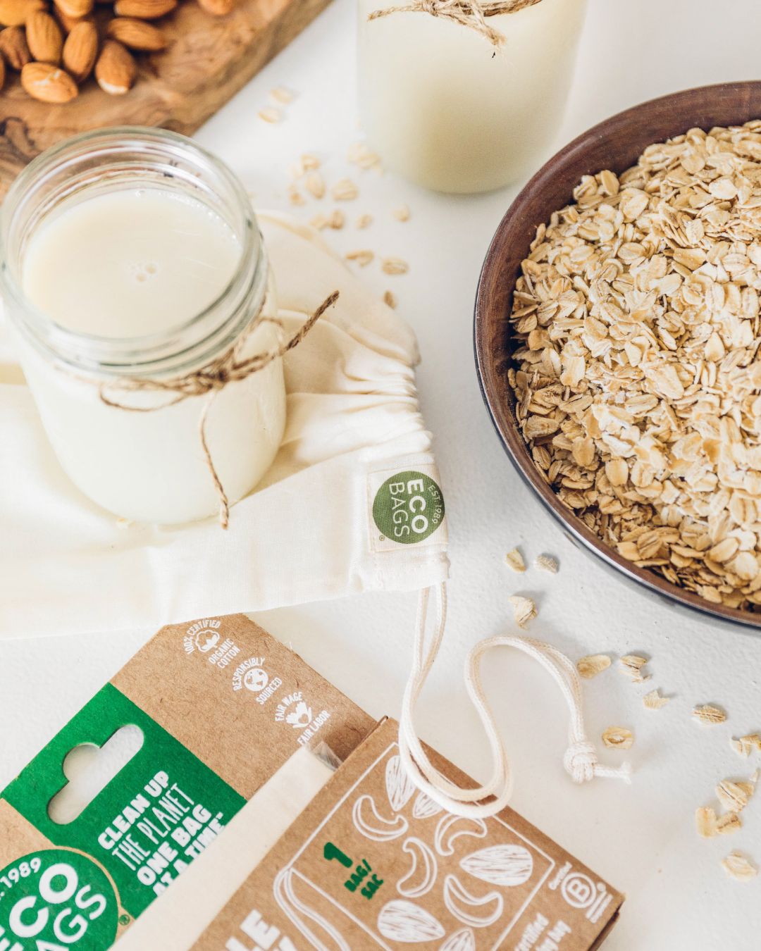 Oatmeal and almond ingredients with a jar of milk on a white surface, featuring 'Eco Bags' packaging.