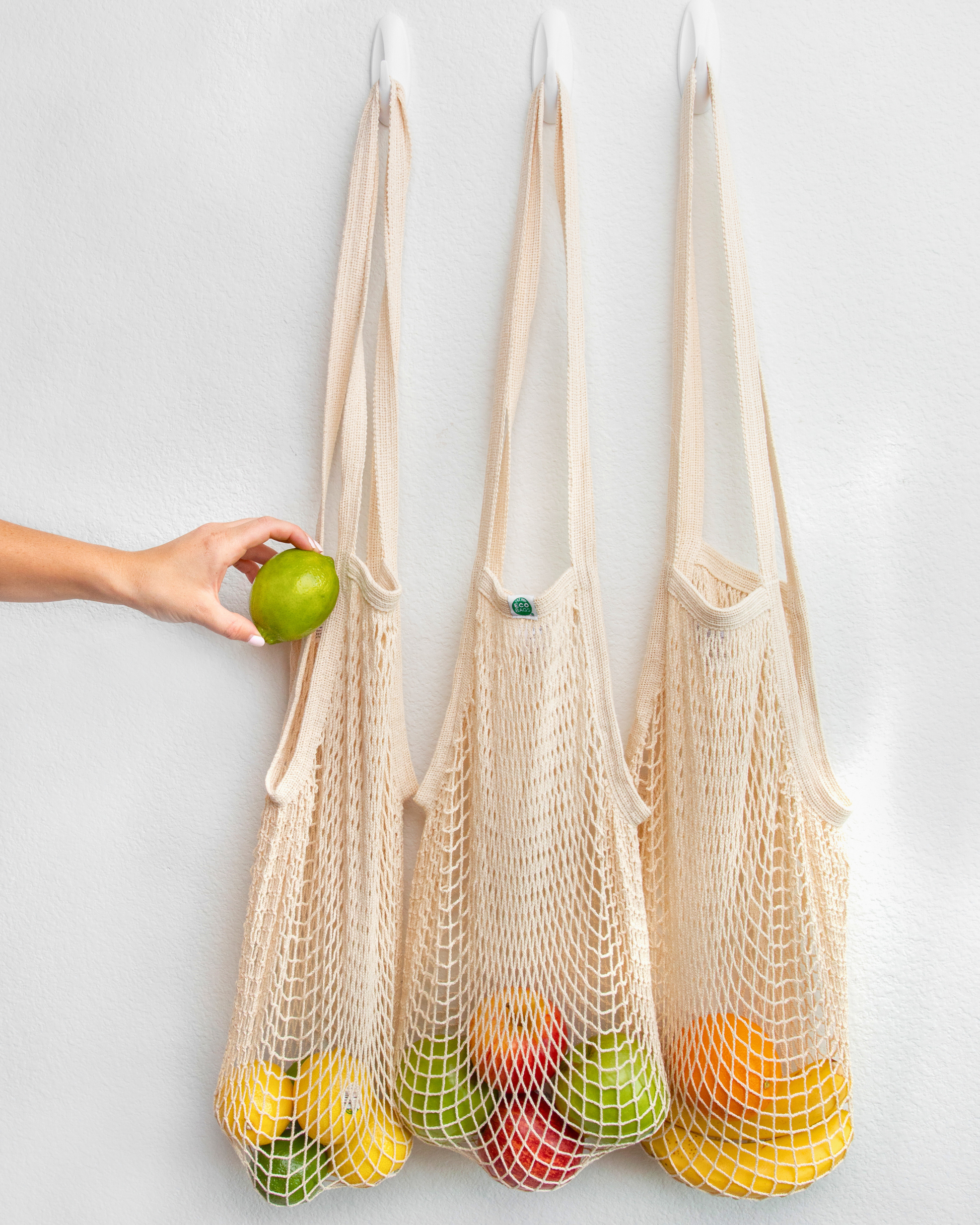 Three beige, natural cotton mesh bags filled with produce against a white background