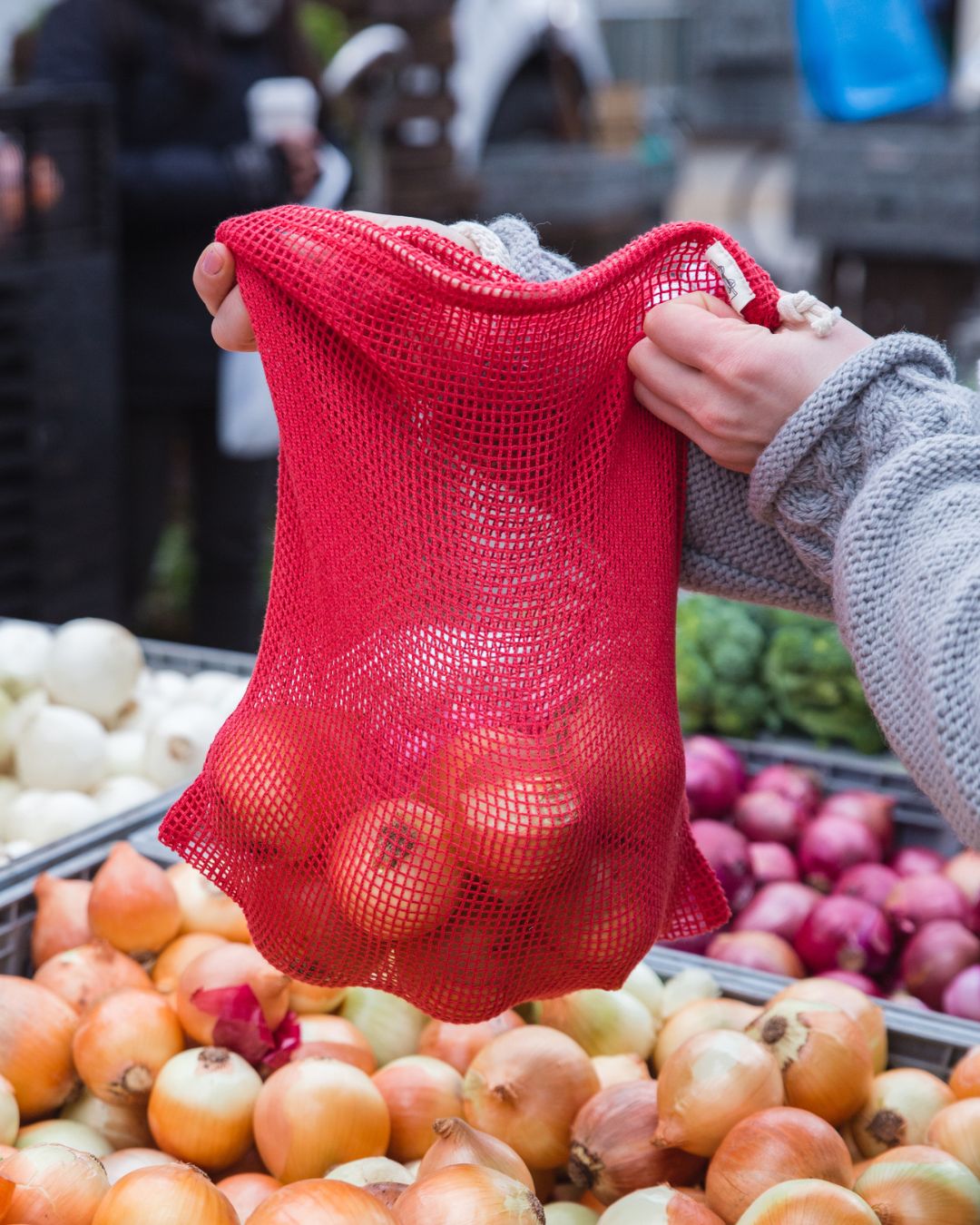 Red mesh bag with apples held over a produce stand with onions and other vegetables.