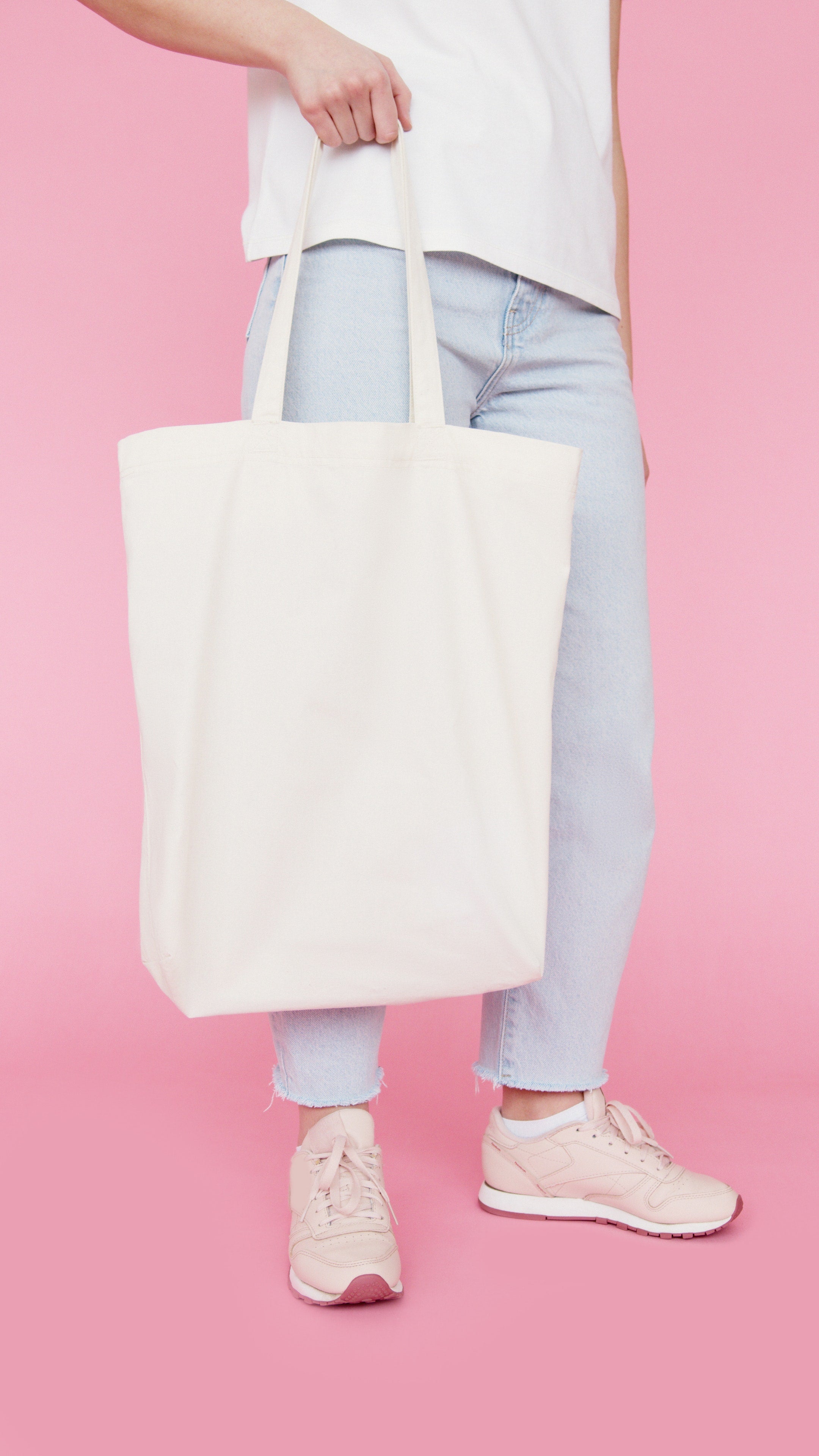 Person holding a natural organic canvas book tote bag, standing against a pink background.