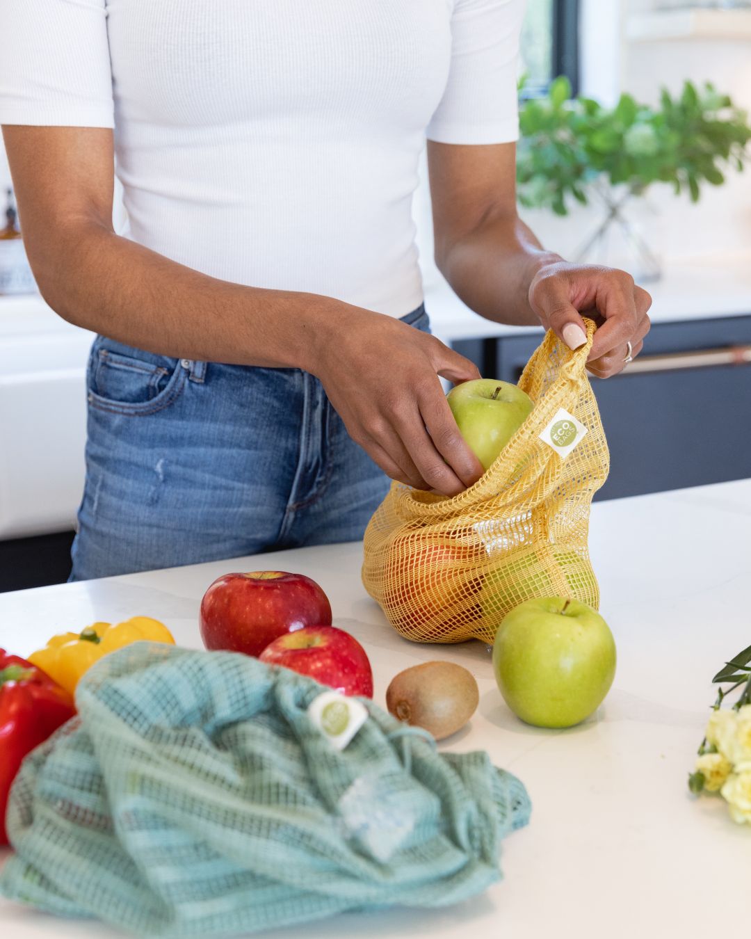 Person using a yellow reusable produce bag with fruits on a kitchen counter.