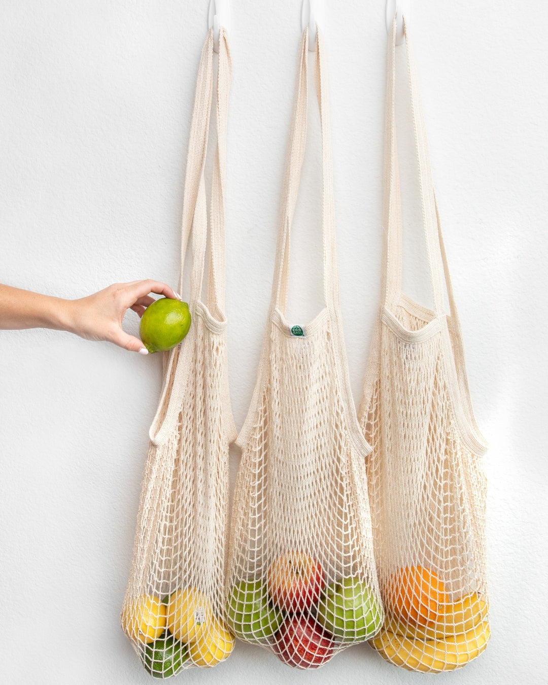 Three natural undyed string bags filled with fruits against a white background