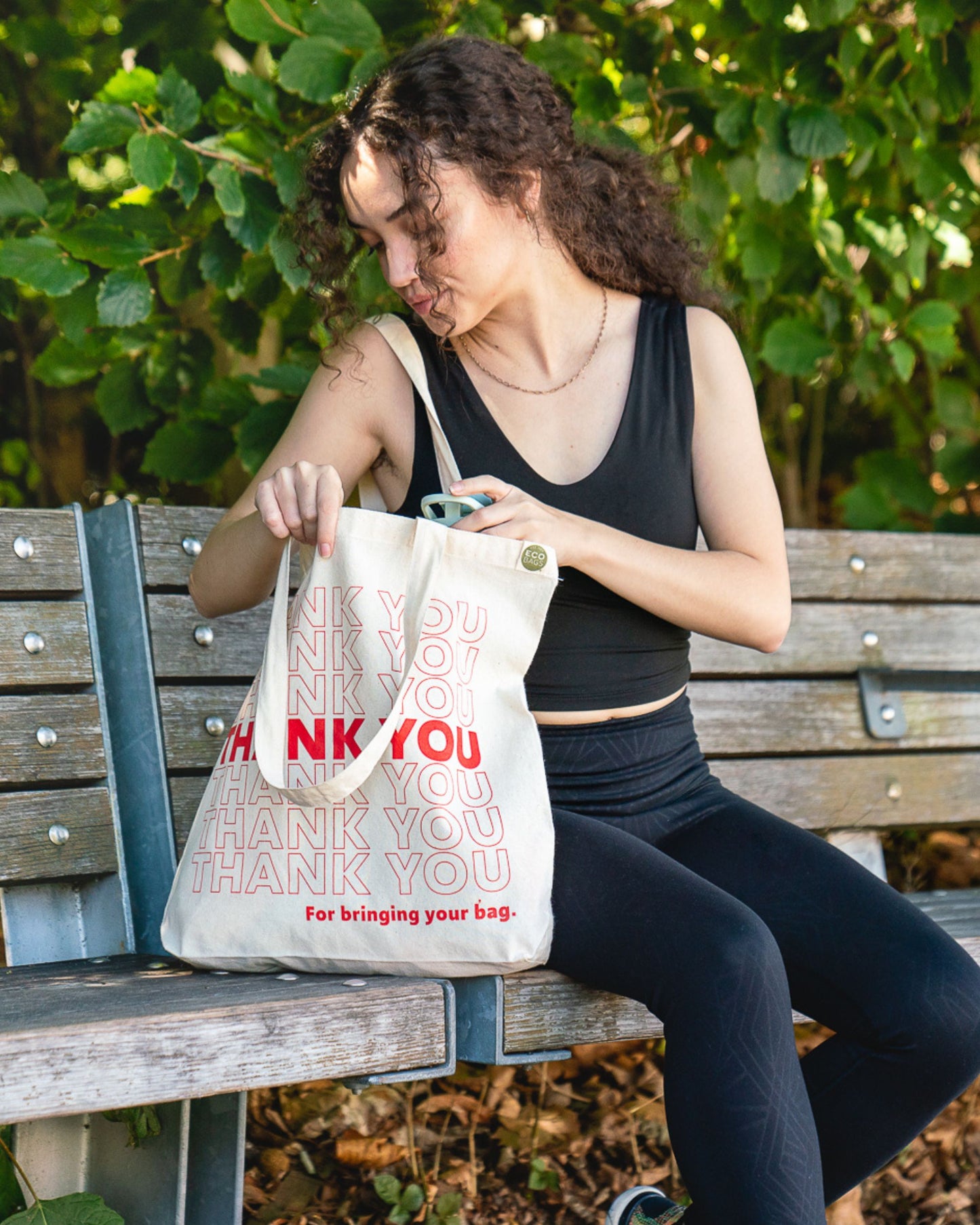 Person sitting on a bench holding a 'THANK YOU' bag with greenery in the background