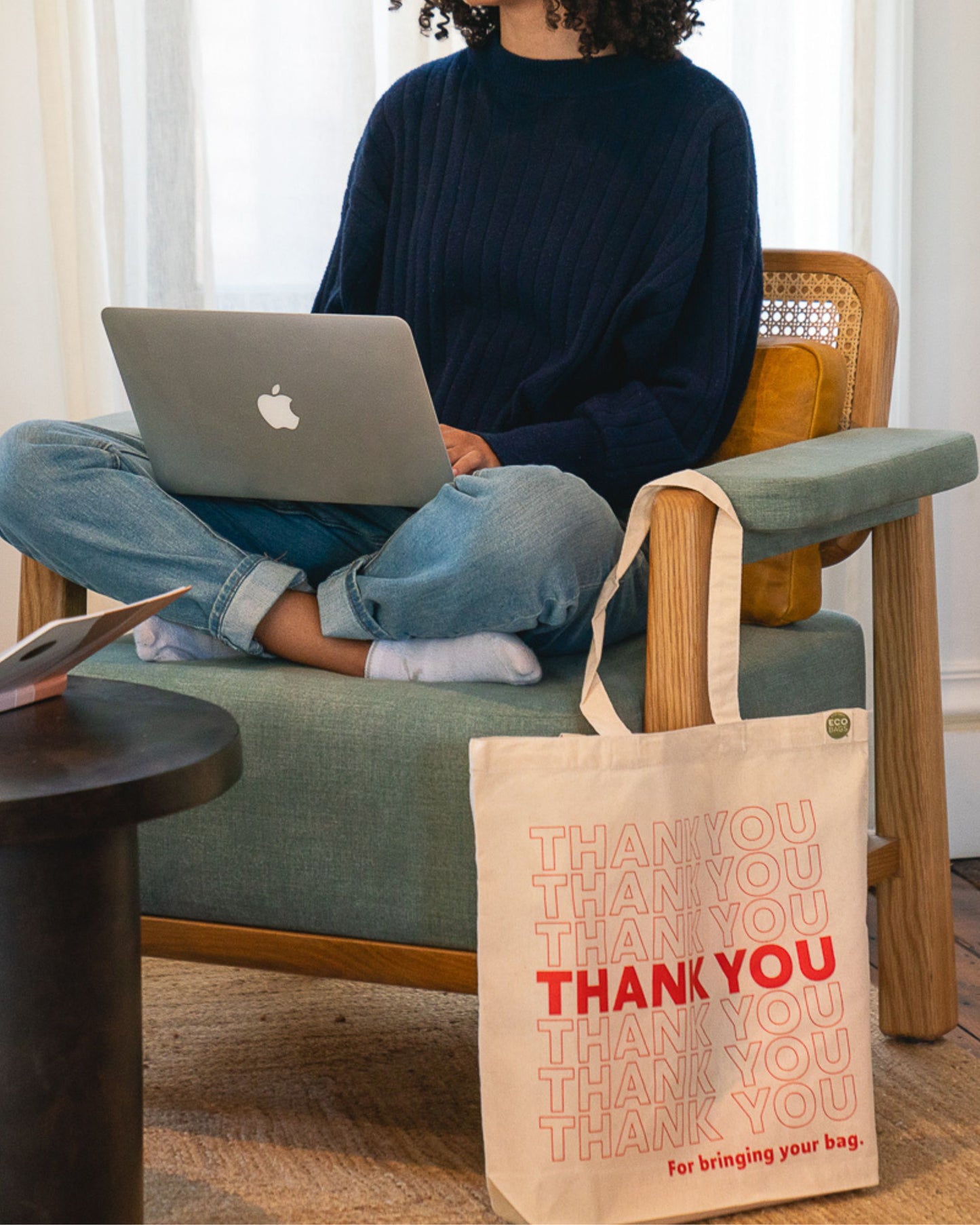 Person sitting on a chair with a laptop, next to a tote bag with 'THANK YOU' text.