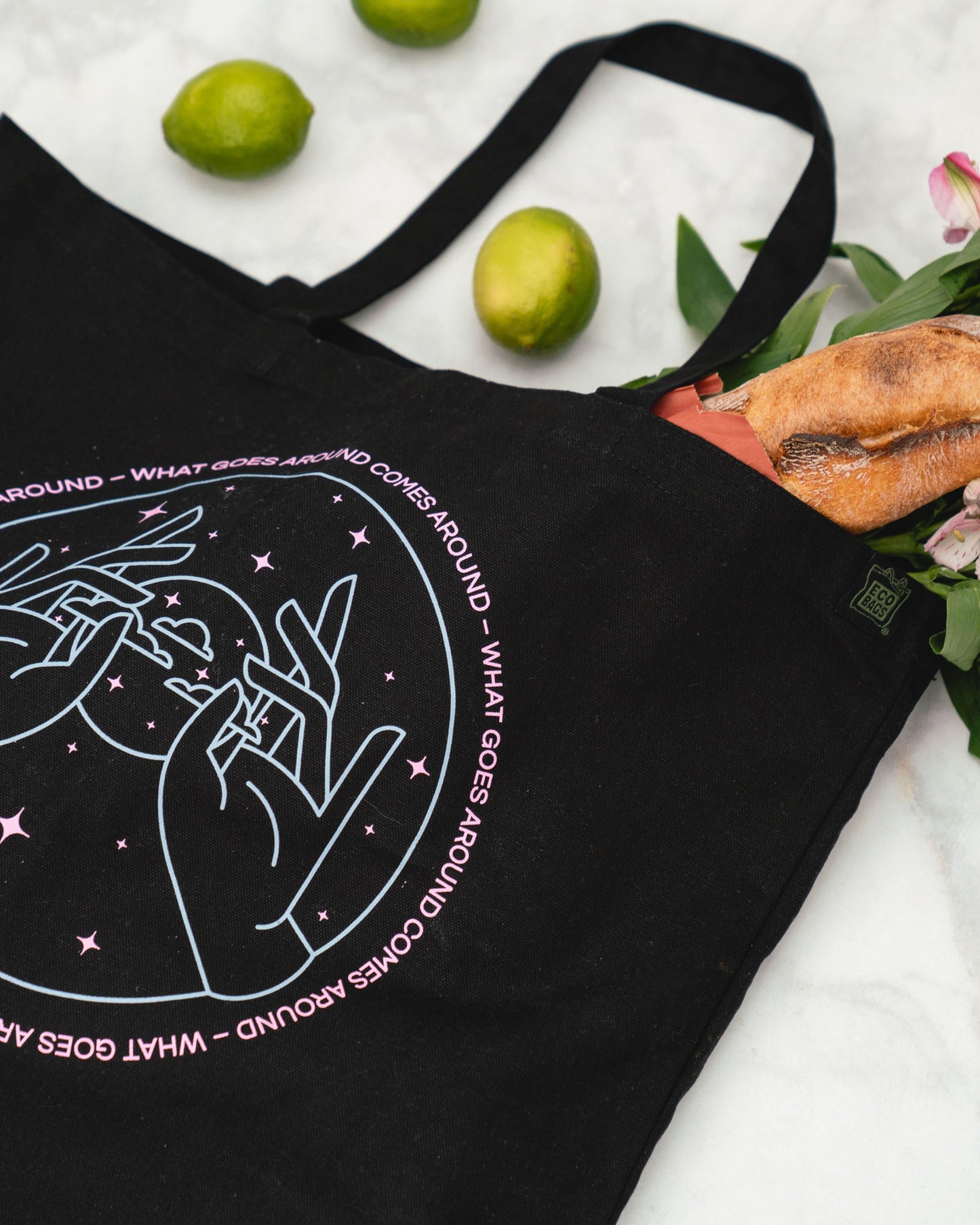 Black tote bag with a pink and blue design and text on a marble surface with limes and flowers.
