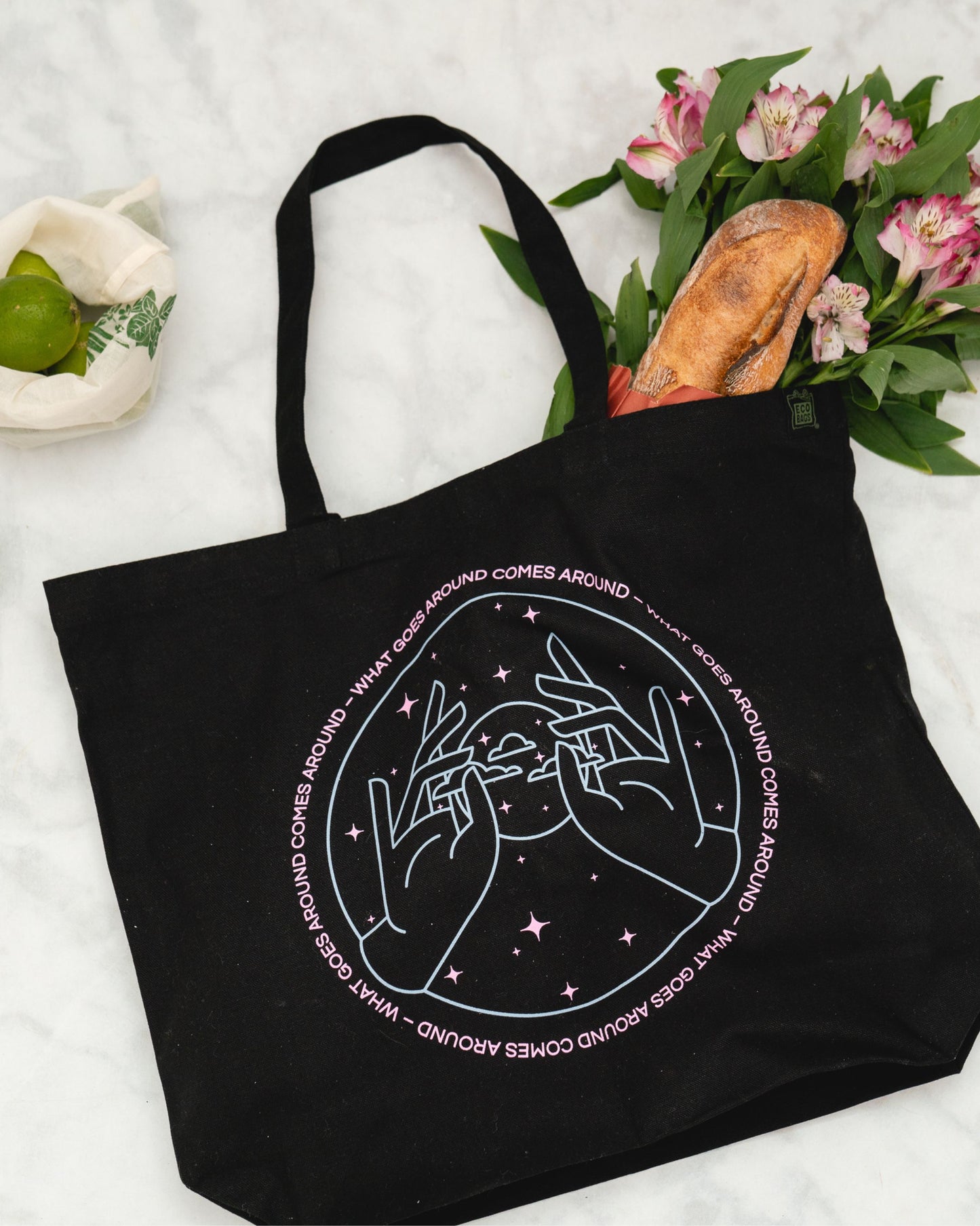 Black tote bag with pink and blue graphic design on a light gray surface with flowers and bread.