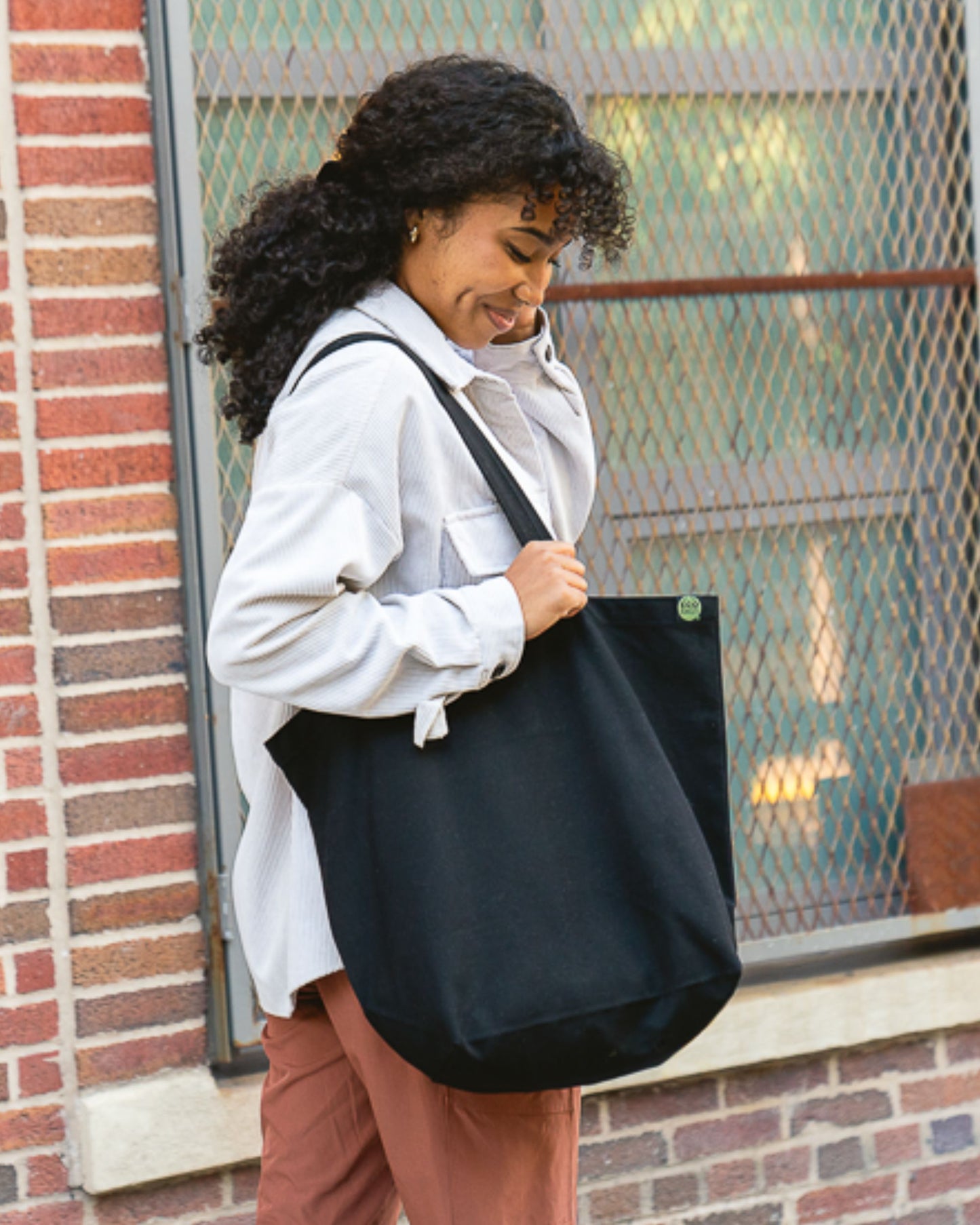 Person with curly dark hair holding a black tote bag against a brick wall.