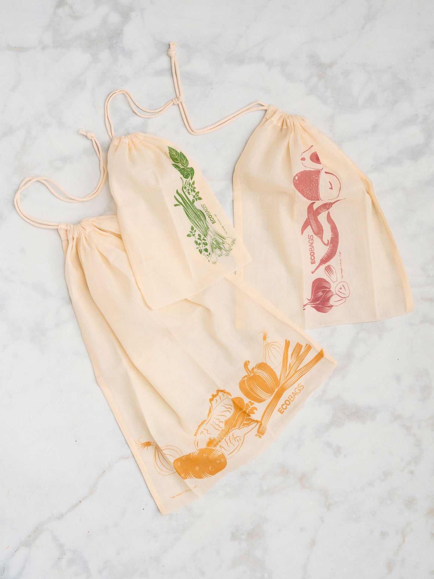 three natural cotton drawstring bags with printed produce graphics on a light background