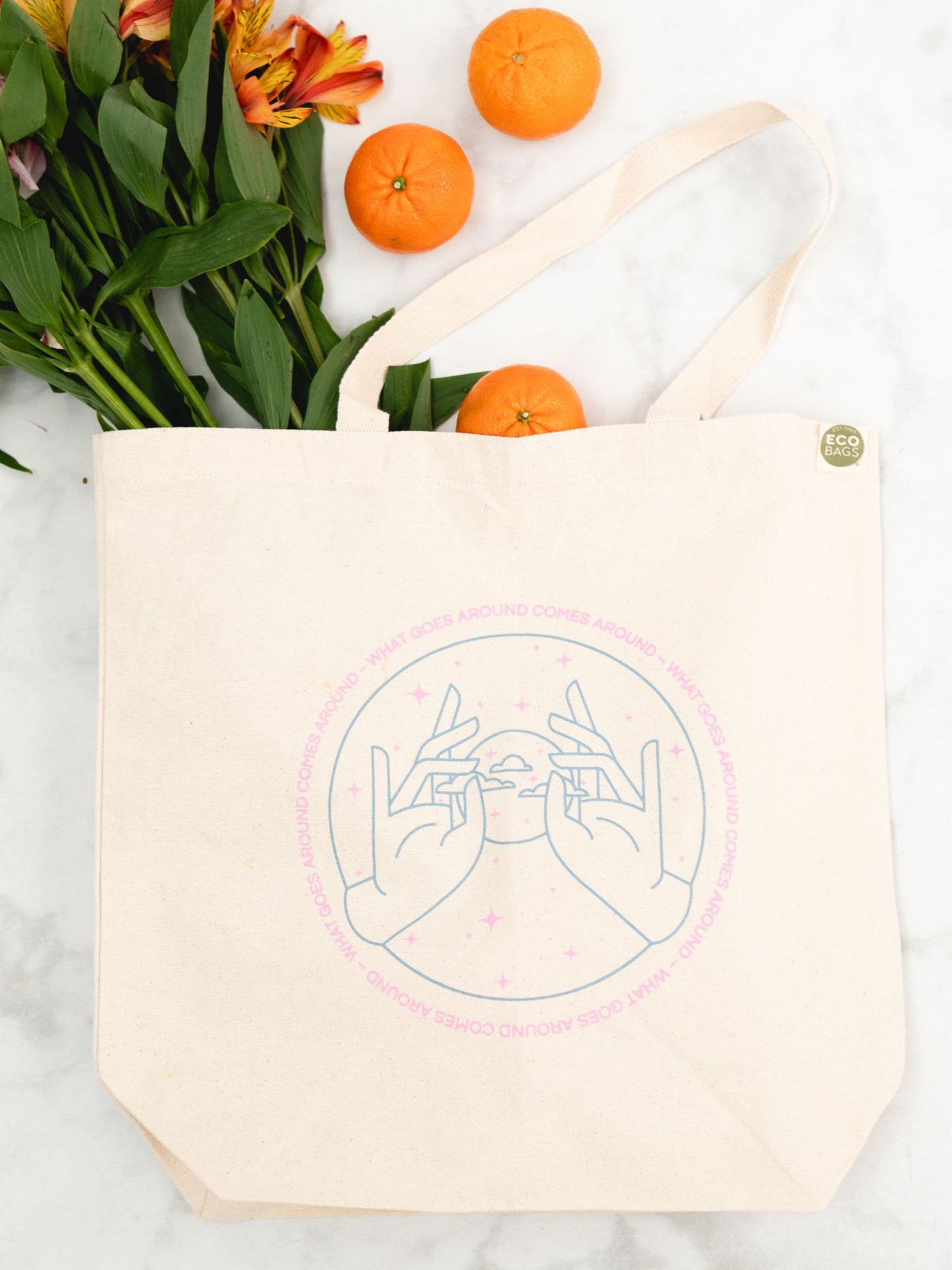 Natural, untreated cotton tote bag with a blue and pink design on a marble surface with flowers and oranges.