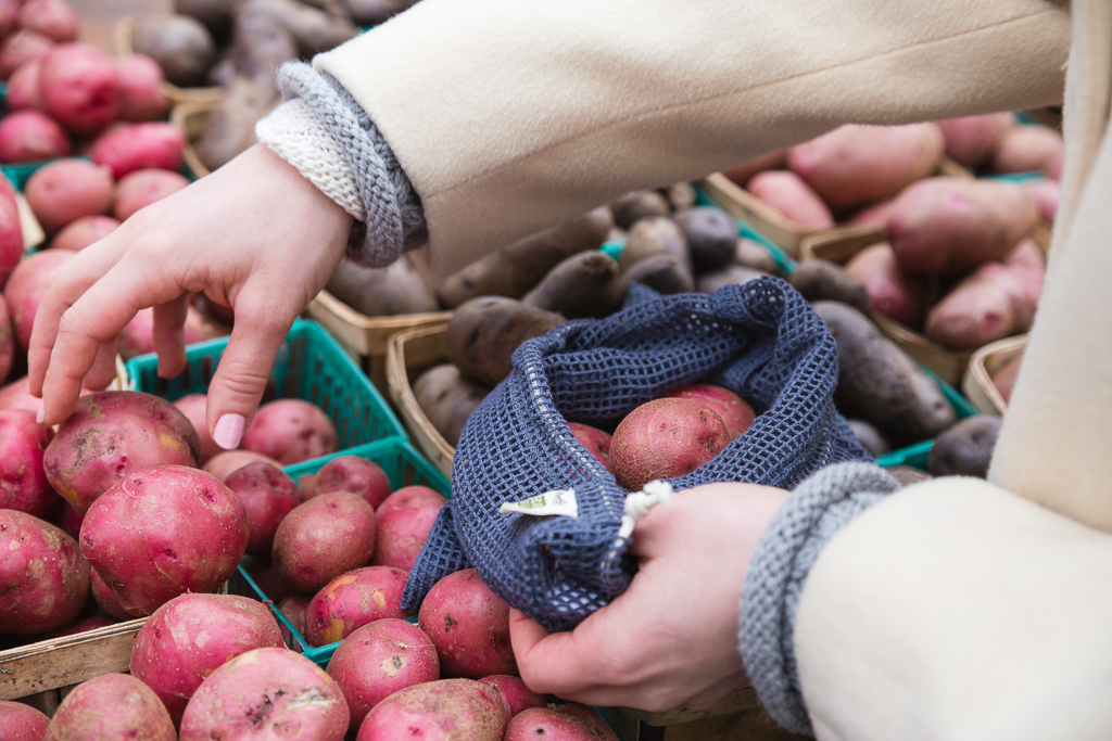 A hand reaches for a small potato while holding a mesh bag full of other produce