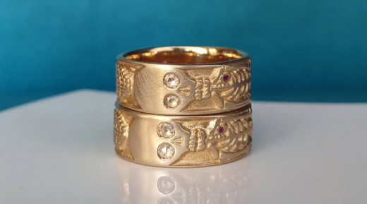 Two shiny gold rings from Bittersweets NY, with skeletons with diamonds for eyes are stacked on top of another on a white countertop with a blue background. 