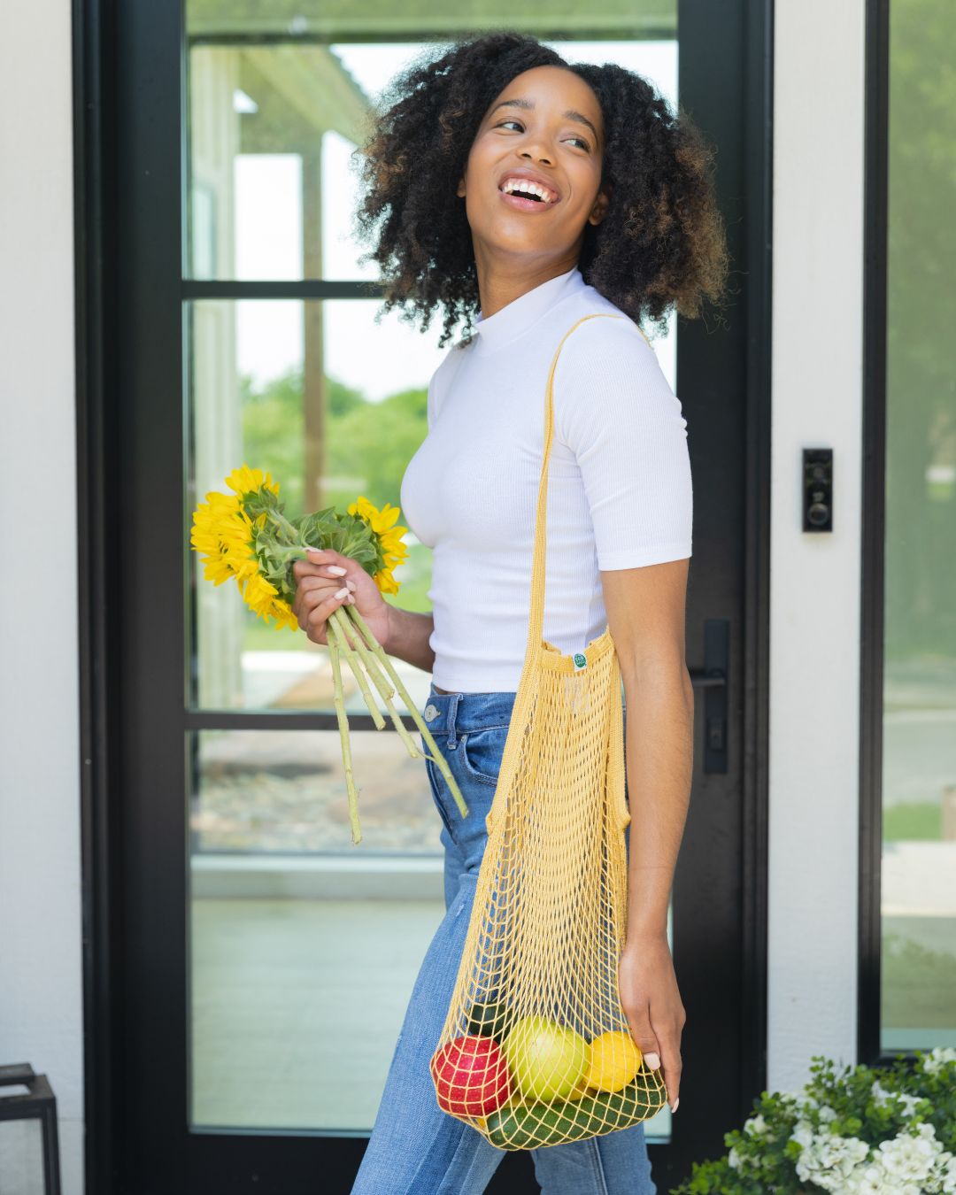 Person holding an eco-friendly yellow reusable bag with groceries and flowers, standing in front of a door.