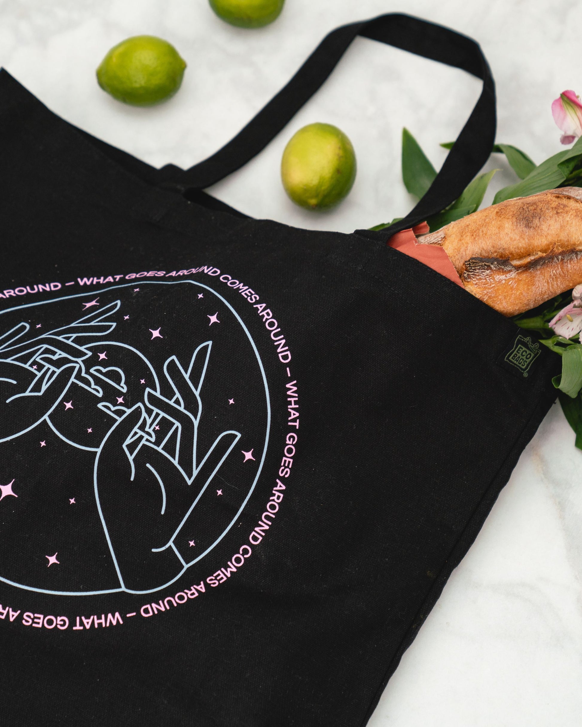 Black tote bag with a pink and blue design and text on a marble surface with limes and flowers.