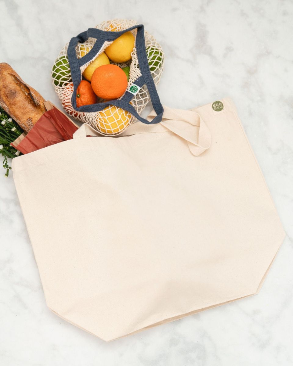 A natural colored reusable tote bag lays against a white marble surface with a baguette and mini string bag beside it